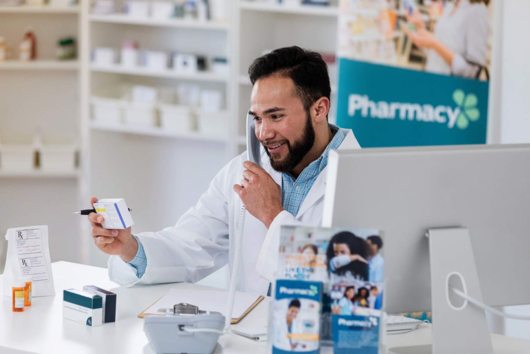 Pharmacist speaking on phone, holding medication box at counter in a modern pharmacy.