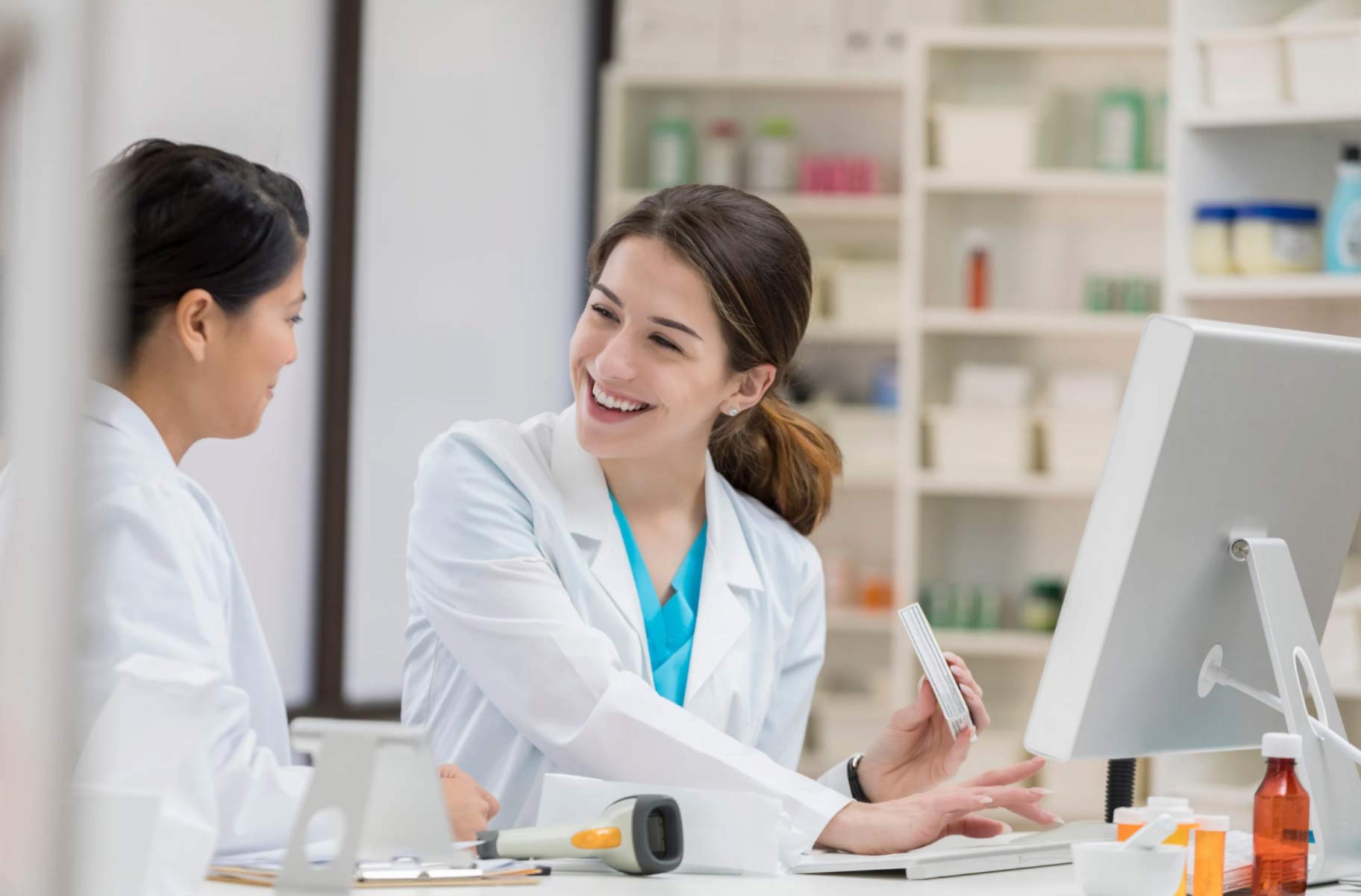 Two pharmacists in lab coats smiling and talking while working at a computer in a pharmacy.