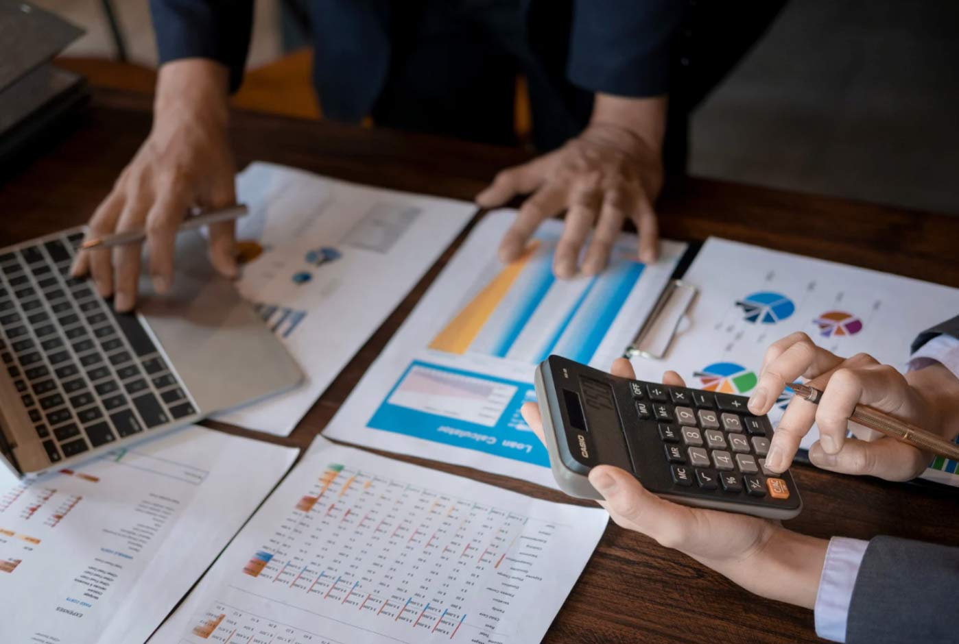 Two people working with charts, a laptop, and a calculator at a desk covered in financial documents.