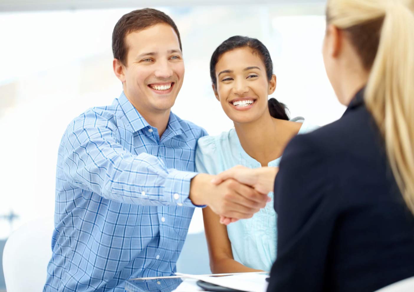 Smiling couple shakes hands with a businesswoman across a desk in a bright office setting.