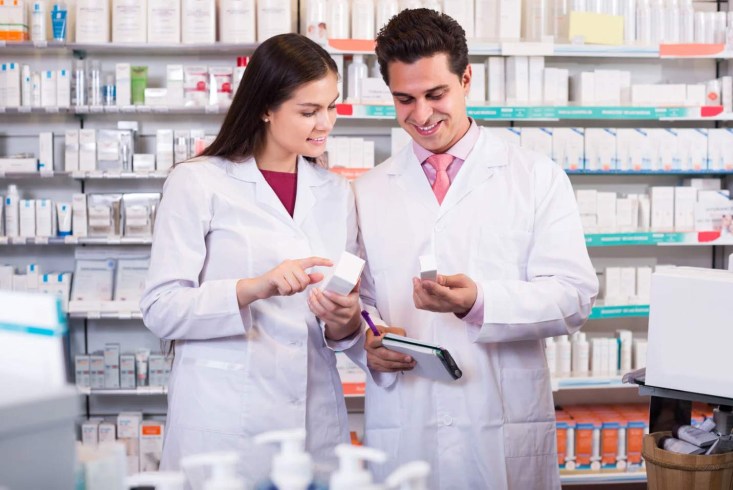 Two pharmacists in white coats discussing medication boxes in a pharmacy aisle.