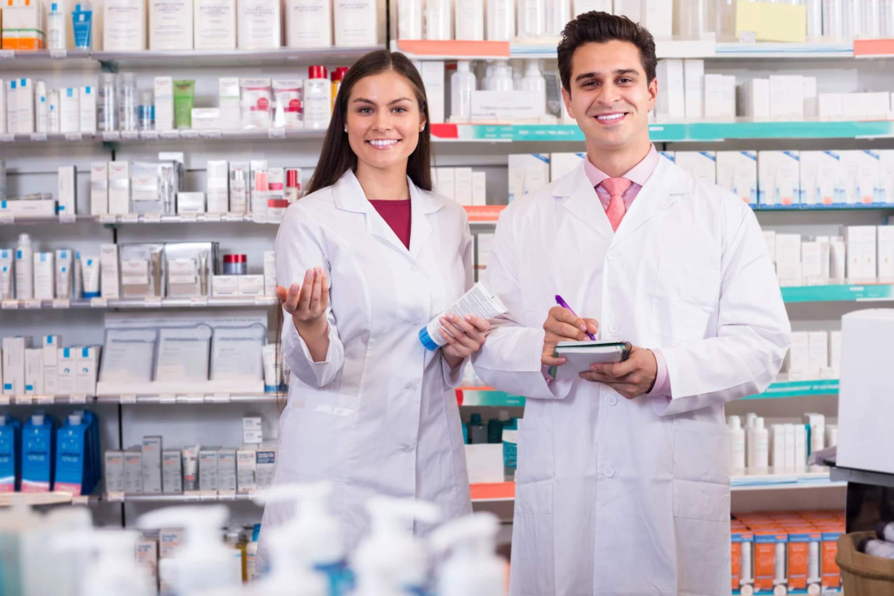 pharma-1 Two pharmacists in white coats stand smiling in a pharmacy, holding a notepad and a product.