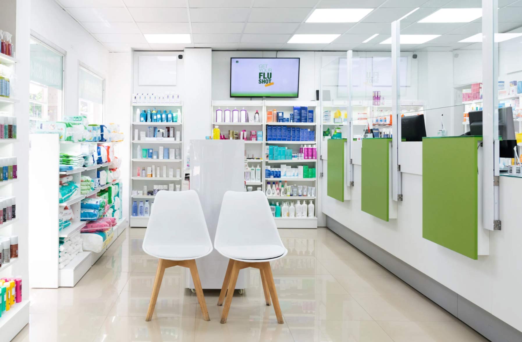 Modern pharmacy interior with shelves of products, white chairs, and green counter partitions.