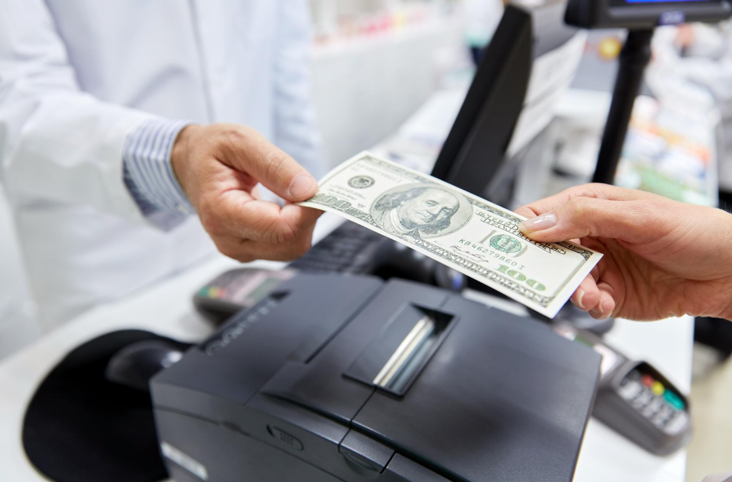 A person hands a $100 bill to another person across a counter with a cash register visible.