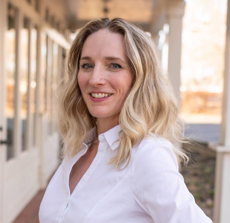 Woman with wavy blonde hair in a white shirt smiles outdoors near a building with large windows.