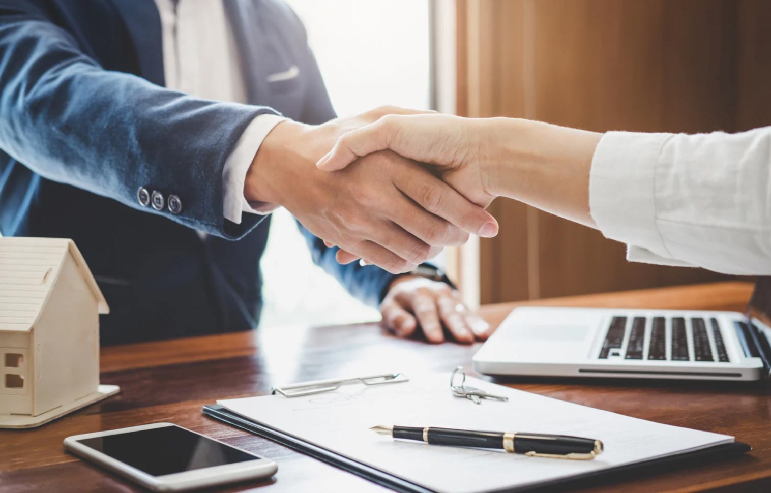 Two people shaking hands over a desk with a laptop, clipboard, phone, and small house model.